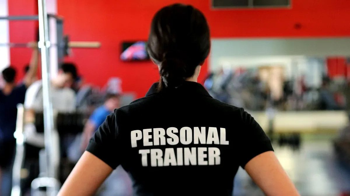 A person wearing a black shirt with "PERSONAL TRAINER" written on the back, standing in a gym with other people exercising in the background.