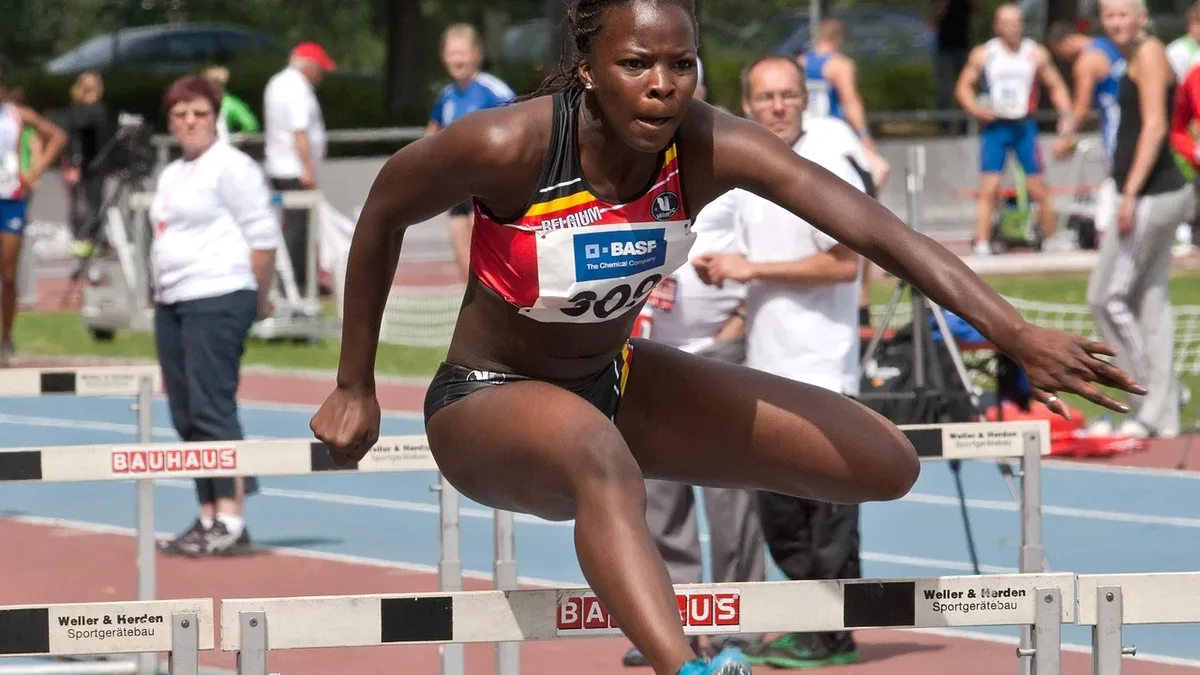 A woman in athletic attire jumping over a hurdle on a track, with other people in the background.